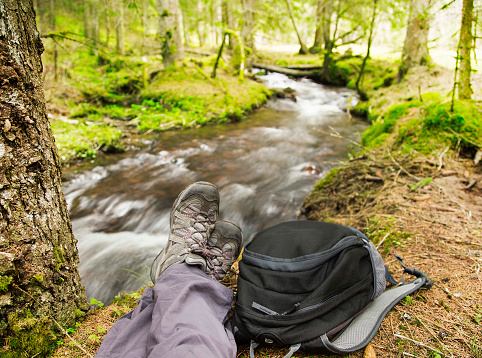 Hiker Legs with Boots and Backpack Resting in the Forest next to the River