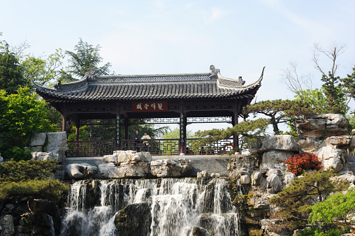 Yangzhou, China - April 21, 2015.  A Chinese couple standing with hands on rail in a pavilion above waterfalls with some other people sitting behind in a scenic public park in Yangzhou. Yangzhou is an ancient city located in the south of Jiangsu Province, China, with a history of over 2,000 years.