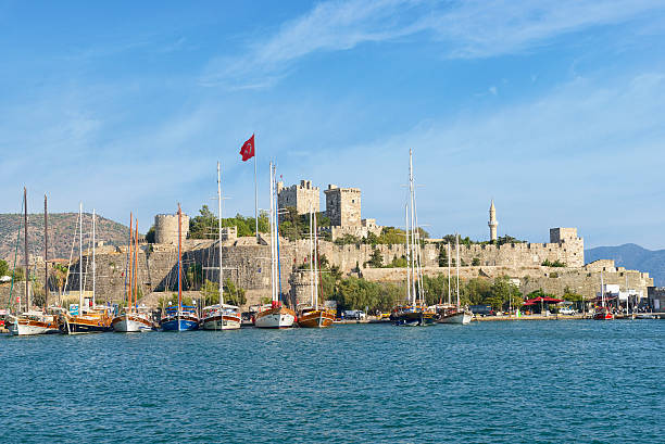 A traditional Turkish gulet sailing boat glides into the popular seaside resort of Bodrum on the Aegean Sea with the Bodrum fortification in the background. Bodrum is considered the St. Tropez of Turkey. Gulets are traditional boats that take visitors on long or short trips along the beautiful coastline. The Turkish flag waves from the gullet and the fort.