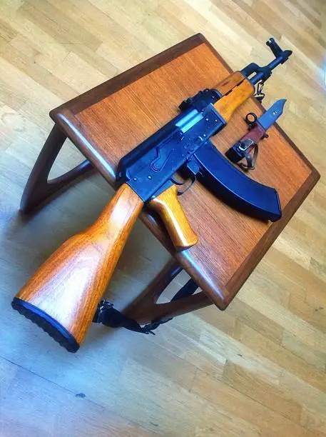 A AK-47 with bayonet knife resting on a British made original 60s G Plan Astro table shot indoors against an oak wood floor.