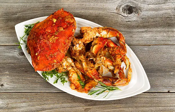 High angled view of freshly cooked crab with spicy sauce and herbs on white serving plate. Rustic wood underneath dish.