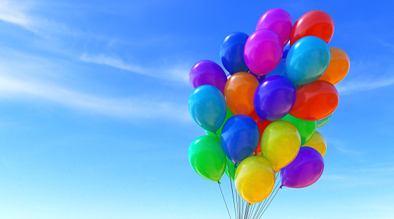 A group of colorful balloons tied together into a bunch, isolated on a soft blue sky background. The balloons surface looks slightly marbled and they come in all colors of the rainbow.