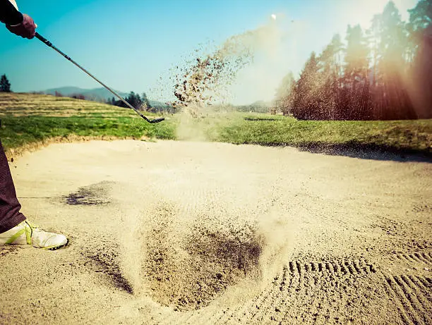 Golfer hitting out of a sand trap. The golf course is on the sand. Sand making splashes. Sun and sunshine in the background