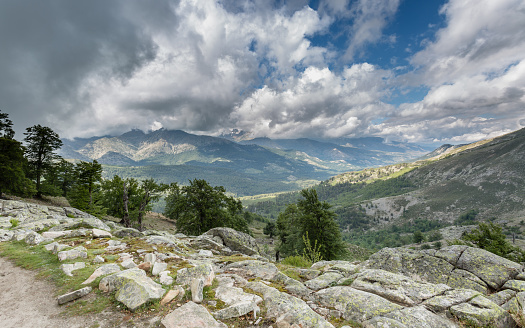 View across Corsican mountains from the GR20 track near to Lac de Nino with clouds and blue sky in the distance and trees and rocks in the foreground