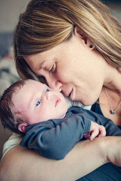 Mother cuddling her newborn baby, close-up on faces. Close-up on the faces of young blonde mother tenderly cuddling her newborn baby. Baby is 10 day's old, has dark hair and open blue eyes. He is wearing a grey onesie. Vertical indoors shot with natural light. unknown gender stock pictures, royalty-free photos & images