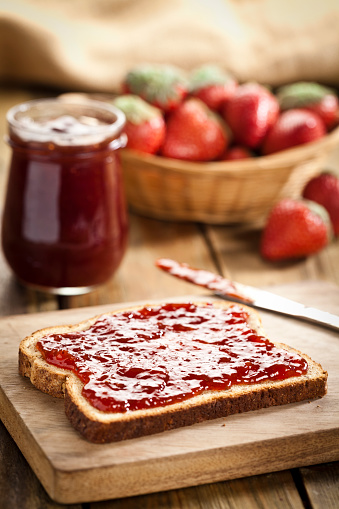 Bread slice spread with marmalade sits on a rustic wooden table with a table knife smeared with jam on right side. Marmalade jar is beside a strawberry basket behind bread slice. Shallow deep of field. Shot with a DSLR Canon EOS 5D Mark II