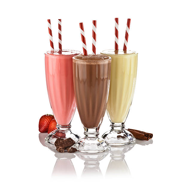 Chocolate, vanilla and strawberry milkshakes shot on reflective white backdrop. The milkshakes are served in classic glass and arranged as a triangle. There are two old-fashioned red-and-white straws standing in each glass. At the base of the glasses are chocolate pieces, a strawberry fruits and cinnamon sticks. Visible reflection on foreground. DSRL studio photo taken with Canon EOS 5D Mk II and Canon EF 70-200mm f/2.8L IS II USM Telephoto Zoom Lens