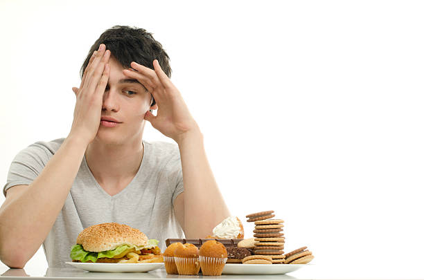 Man having in front sweet dessert and a big hamburger. Young man with chocolate, cupcakes, biscuits and a burger in front not wanting to look and eat. Trying to get fat eating fast food, junk food and lots of sugar. biscuit quick bread stock pictures, royalty-free photos & images