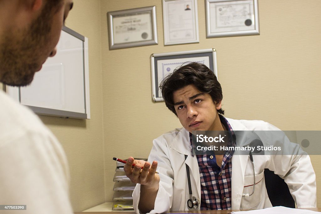 Sad young male doctor consoling his pattient Sad young male doctor consoling his pattient. Pattient looking down with a sadness expression,his eyes closed. They are doctor's office. There is report on the table. Doctor wearing lab coat and holding a pencil. Horizontal composition. Image taken indoors and developed from Raw format. 20-29 Years Stock Photo