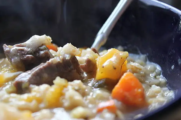 This image shows a beef stew with barley, chunks of beef, butternut squash, carrots served in a dark blue bowl. This is part 2 of 3-part series. One of the other images shows a wider shot with the edge of bowl and the wooden background showing. The other image is a slightly wider take of the stew, with more of the bowl showing, 