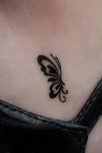 Very small black butterfly tattoo on woman's ankle just above sandal strap of her shoe