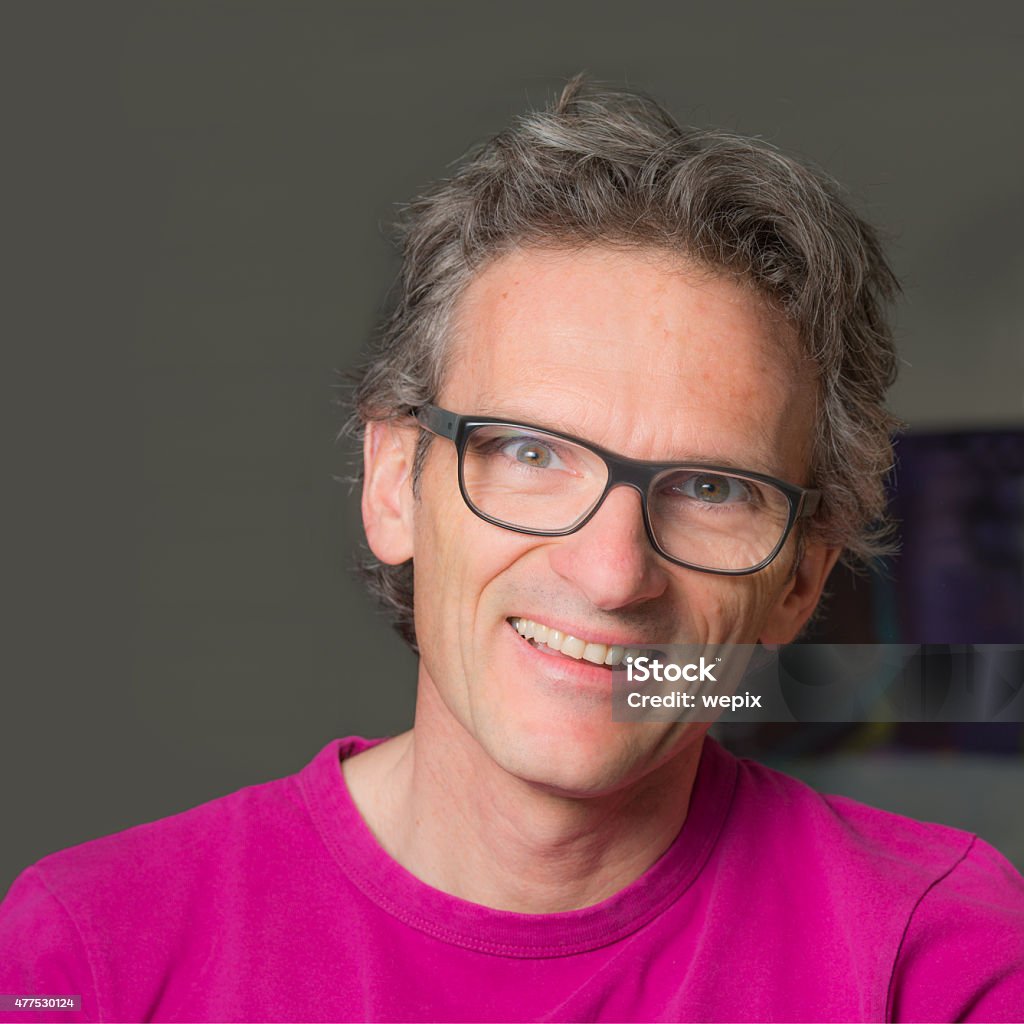 Smiling interesting nice mature casual man glasses best age looking Portrait of a serene man with an open toothy smile who looks into the camera with bright eyes. Real people, the image was taken at his home. He is casually dressed, wears black rimmed glasses and a pink t-shirt. His brown hair has some gray streaks. 2015 Stock Photo