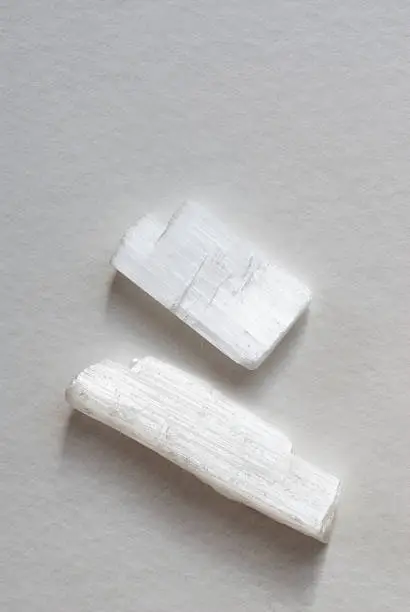 Two rough cut pieces of selenite, which is a variety of gypsum.  They are studio shot on a gray background.  The selenite pieces are white and rectangular.