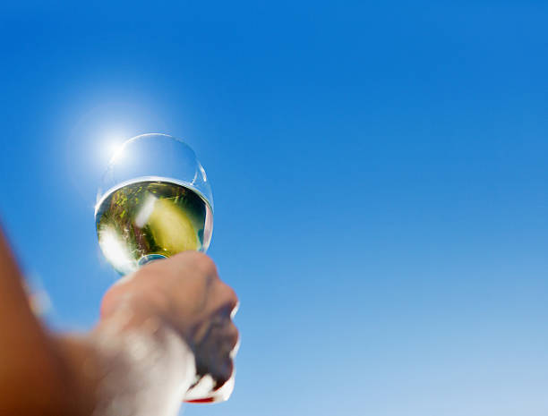 A feminine hand holds a glass of white wine up against a clear blue sky so that the sun seems to be balanced on the rim of the wineglass. Copy space on the sky.