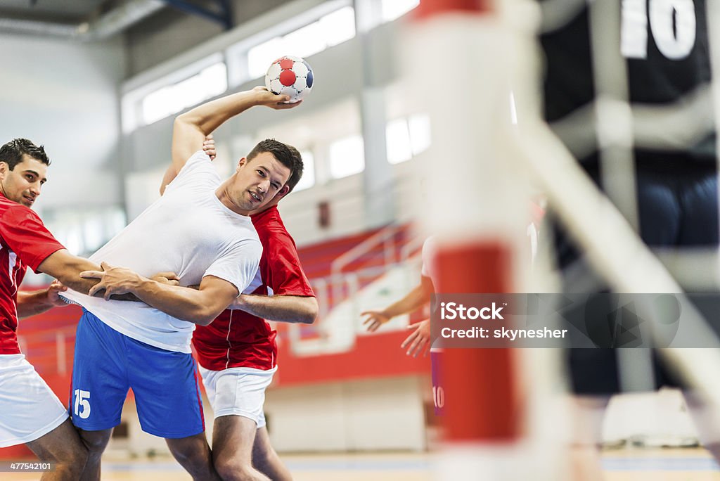 Handball player shooting at goal. Male handball player is shooting at a goal, while his opponents are holding him.    Team Handball Stock Photo