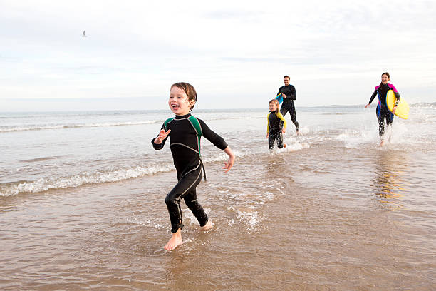 Family in Wetsuits Family of four are running along the shore with body boards. They are all laughing. wetsuit stock pictures, royalty-free photos & images