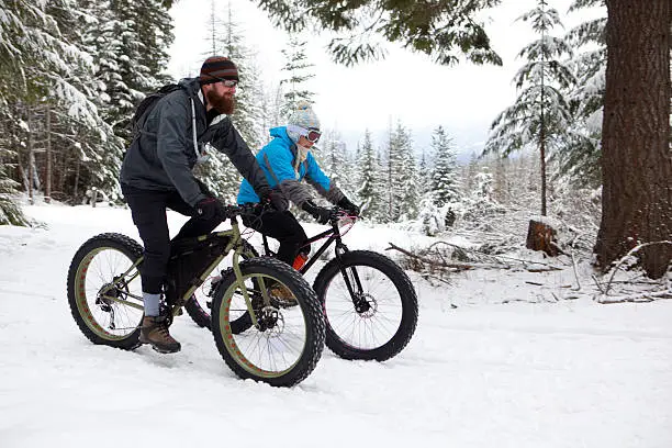 A couple are out for a ride on their fatbikes. This snow covered trail runs through the woods with a nice view of the valley.