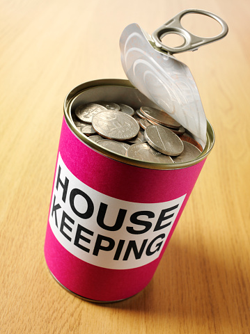 Pink label on an open tin can with the word house keeping filled with American currency on a wooden desk