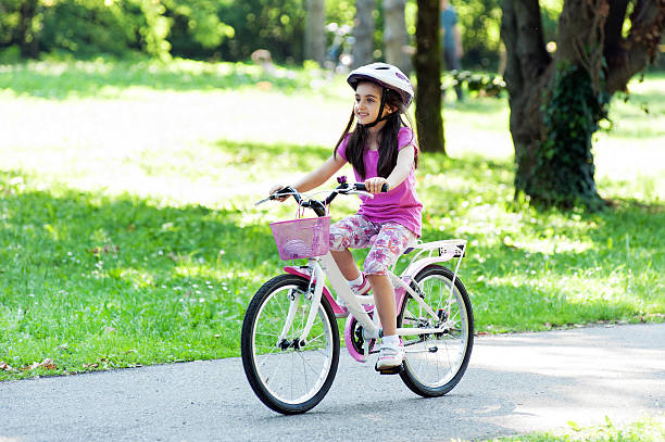 Little girl in a trendy pink outfit with matching pink bike and helmet riding her bicycle along a lane in a wooded park