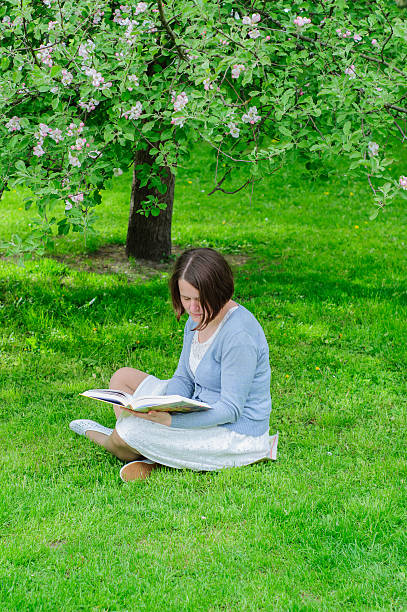 istock Young woman reads a book under blooming tree 477555728