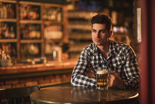 Young man sitting alone at the table in a pub and holding a glass of beer while thinking and looking away.