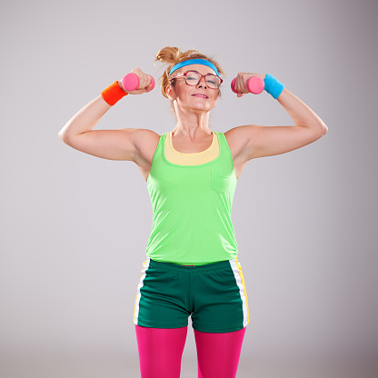 Studio portrait of funny strong fitness female lifting up small pink weights. She is really happy and satisfied young girl. Woman is looking to camera. She is showing us her fit body and biceps muscles. Nerdy young woman with glasses is wearing turquoise headband, light green athletic shirt and dark green shorts. 