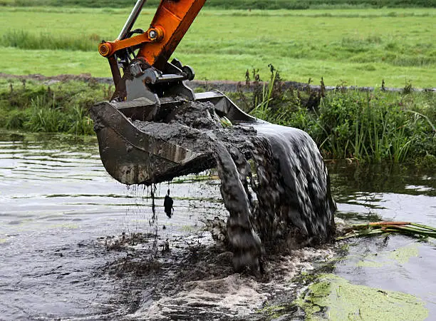 A crane with backhoe takes a scoop of sludge from a canal, part of dredging activities for canal maintenance