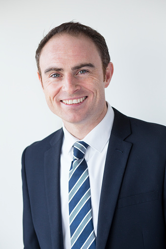 A young male is smiling and looking at the camera, wearing business formal as a businessman. He is wearing a blue blazer, black vest and blue tie. He is in a studio with a isolated white background.