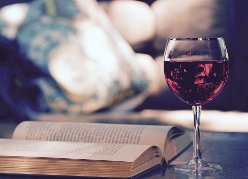 A glass of red wine and a book resting on a glass table outside by a lounge chair in the summer air. Shot in the afternoon.