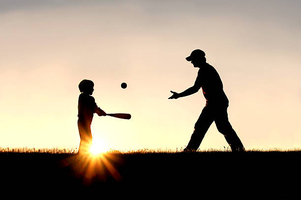 Silhouette of Father and Son Playing Baseball Outside A silhouette of a father and his young child playing baseball outside, isolated against the sunsetting sky on a summer day. baseball sport stock pictures, royalty-free photos & images