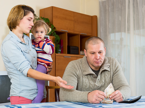 Wife nags his husband about money. She is holding their daughter on her arms he is sitting at the table with cash in his hands indoors