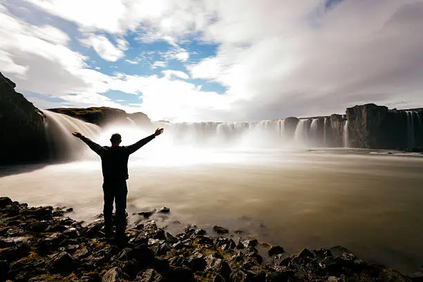 Godafoss waterfall in winter, Skjalfandafljot River, North-Central Iceland. Motion blurred, waterfall, horizontal composition. Long exposure. A man stands looking at the view, arms raised, silhouetted. 