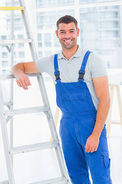 istock Happy handyman in overalls leaning on ladder at office 477649820