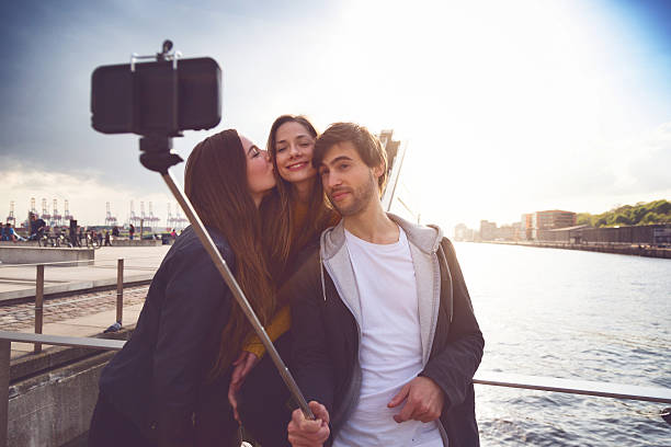 Friends taking a selfie stick in Hamburg. Three friends, two girls and a boy, enjoy with a selfie stick and a smartphone by the Elbe river in Hamburg, taking a self portrait. The girl kiss on her cheek the other girl.
