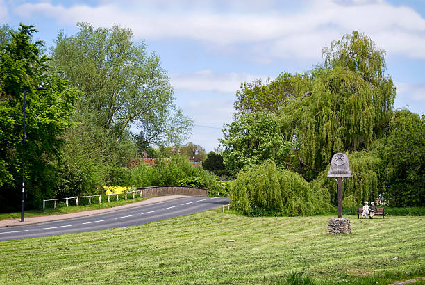 Long Melford, Suffolk, England - May 15, 2015: A man and a woman sitting on a bench near to the village sign and the bridge over the River Stour at Long Melford, Suffolk, in eastern England. Long Melford is - as the name suggests - a very long village. It is also an extremely popular tourist destination.