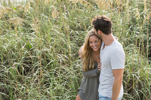 A young, romantic couple walking side by side, arms around each other, on the beach, along the sand dunes.  He is wearing a shirt and shorts, and she has on a beach coverup.  The man is kissing his girlfriend on the top of her head.