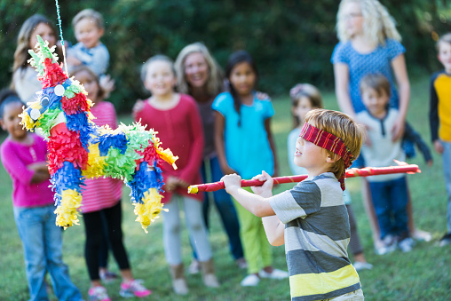 A little boy, 6 years old, tries to hit a colorful pinata with a red stick at a birthday party or cinco de mayo celebration.  He is blindfolded.  A group of mothers and children watch in the background.
