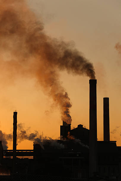 Backlit industrial chimney stacks factory dark smoke pollution rising upwards stock photo