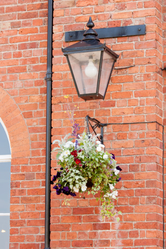 Brightly colored pansies make a lovely show hanging in basket against old brick wall with an old fashioned lamp above them to illuminate it at night