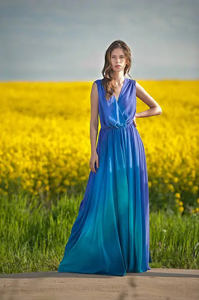 Fashion beautiful young woman in blue dress posing outdoor with cloudy dramatic sky in background. Attractive long hair brunette girl with elegant dress posing in canola field during sunset.