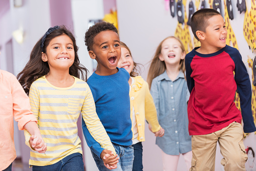 A group of multiracial school children walking through a hallway holding hands, smiling, laughing and shouting.  They are excited to be back to school.  They are preschoolers or kindergarden students, 4 to 6 years old.