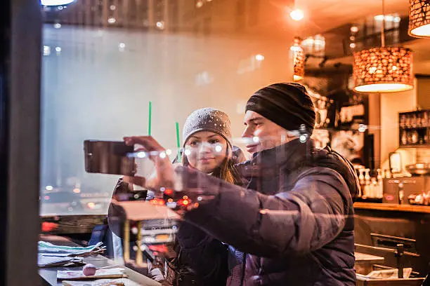 Young tourists, couple, man and woman, make selfie with the smartphone in the cafe in Manhattan. View trough the window. High ISO, noise, defocused photo with reflections.