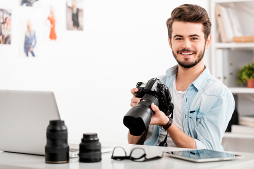 Cheerful young man holding digital camera and smiling while sitting at his working place