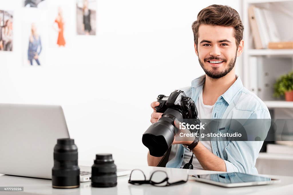 COnfident photographer. Cheerful young man holding digital camera and smiling while sitting at his working place Photographer Stock Photo