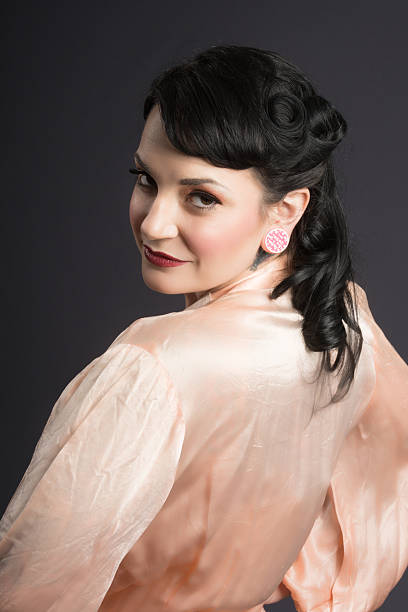 Mid adult beauty in vintage robe, looking over shoulder. Vertical studio shot on gray of smiling woman in hair and makeup inspired by 1940s movies, wearing vintage satin dressing gown. 40s pin up girls stock pictures, royalty-free photos & images