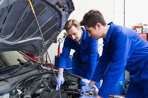 Trainee mechanic looking under the hood of a car with his mentor.