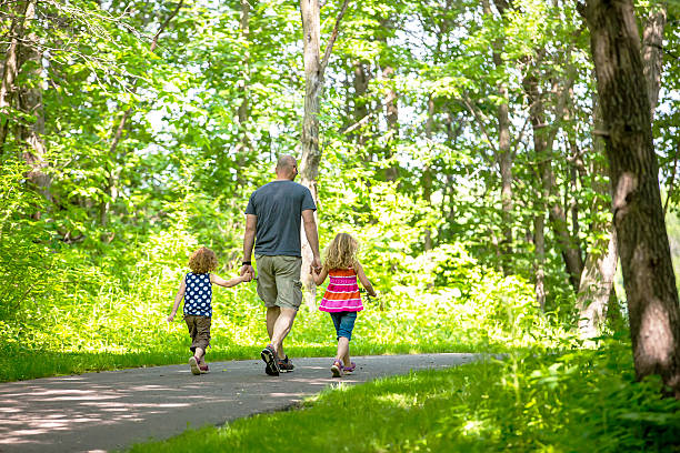 Rear/side view of two young girls going for a walk with their dad on a paved trail at a park. The three are walking side by side and holding hands. It is a beautiful summer day with sunlight hitting parts of the trees and trails.