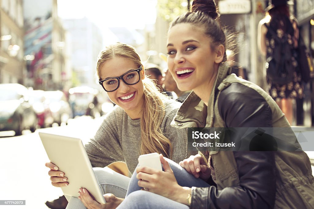 Young women in the city Two young women sitting on the city street, holding a digital tablet and smart phone in hands and smiling at camera. Close-up Stock Photo