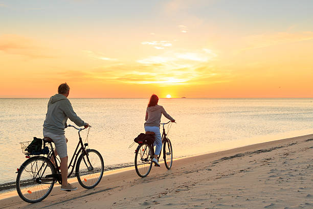 bicicletas de equitação casal maduro ao ar livre na praia ao pôr do sol - action mature adult bicycle senior couple imagens e fotografias de stock