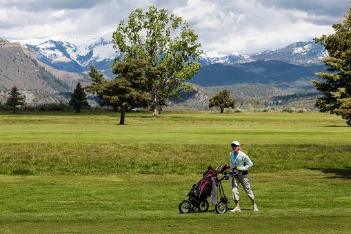 A woman dressed in white walks her clubs on a golf course in Durango, Colorado.   Behind the grassy golf course are snow covered mountains.  The woman appears to be walking briskly pushing her golf clubs with a three wheeled push cart.